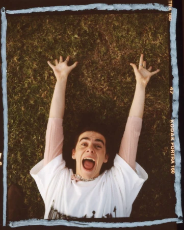 Young adult lies on grassy lawn with arms raised and mouth open, wearing a white graphic tee and beaded necklace, shot in daylight.
