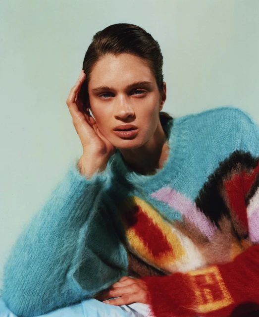 Portrait of a young adult female with dark hair, pale skin, wearing a colorful oversized knit sweater against a pale background.