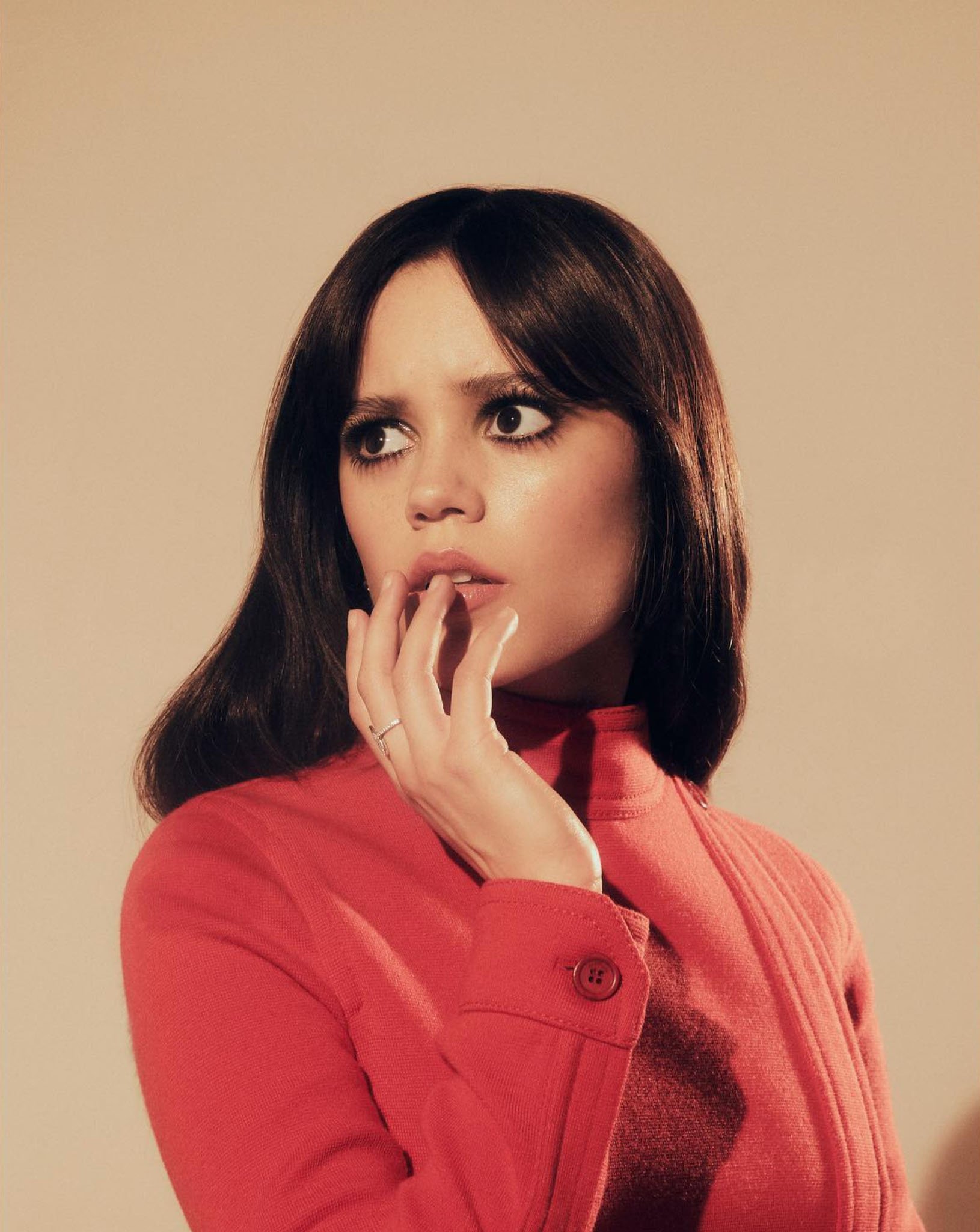 Jenna Ortega wearing a red turtleneck top with dark eye makeup against a warm neutral background.