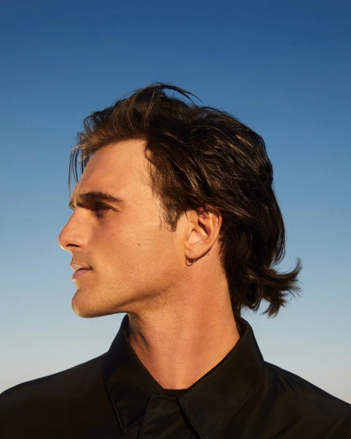 Profile shot of a young adult male with brown hair wearing a black collared shirt against a clear blue sky backdrop outdoors.