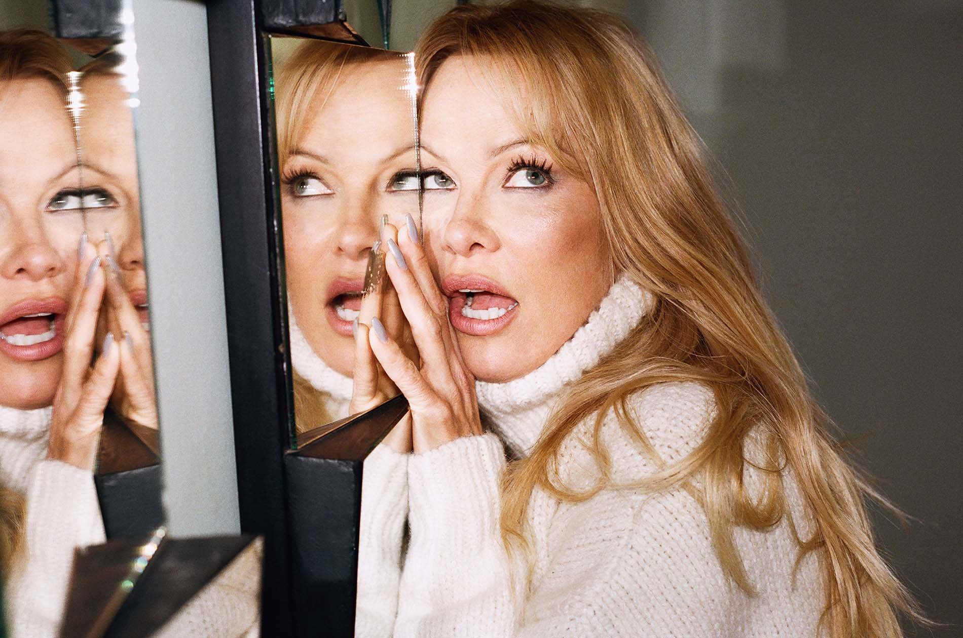 Adult woman with blonde hair wearing a white turtleneck sweater posing near a shattered glass mirror, close-up portrait.