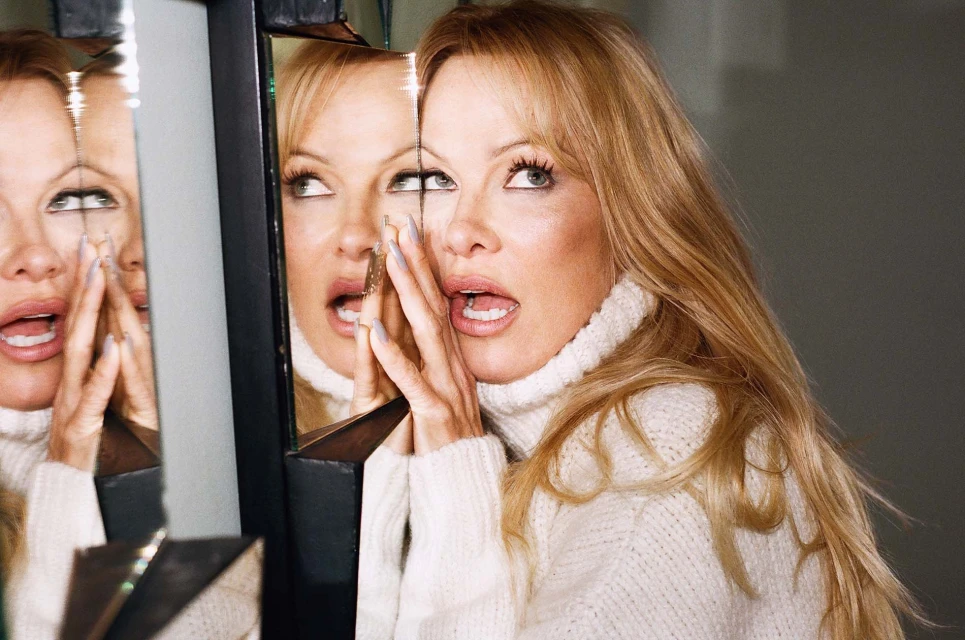 Adult woman with blonde hair wearing a white turtleneck sweater posing near a shattered glass mirror, close-up portrait.
