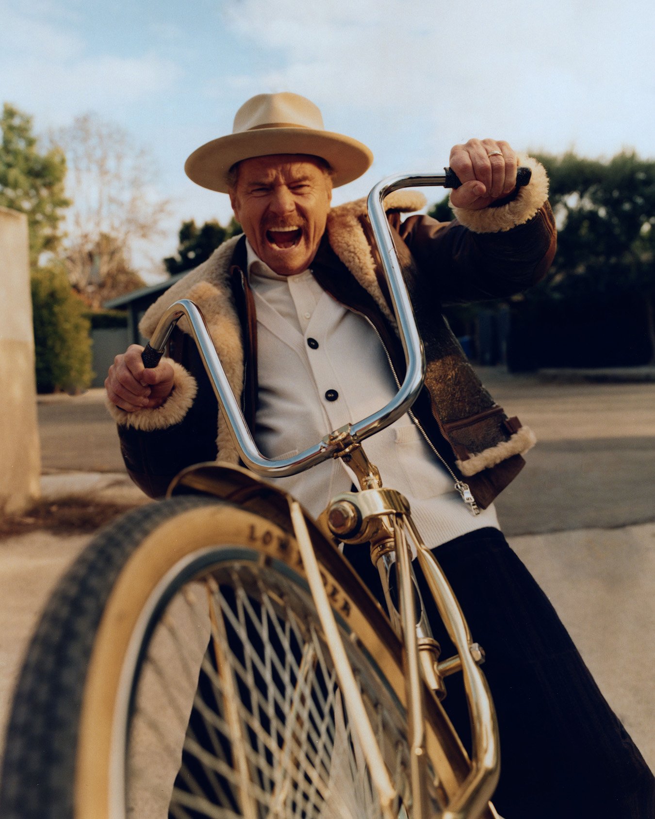 Older adult man riding a bicycle outside, wearing a hat and jacket with fur trim, making an excited expression in natural light.
