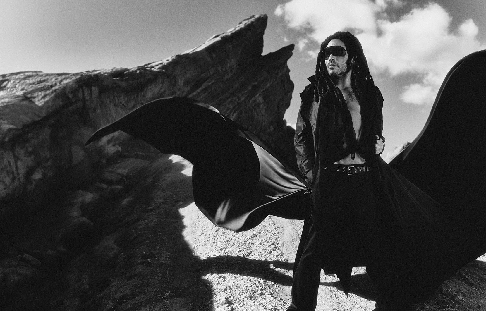 Monochrome outdoor portrait of a dark-haired adult male with dreadlocks wearing sunglasses, a long black coat, standing on a rocky landscape.