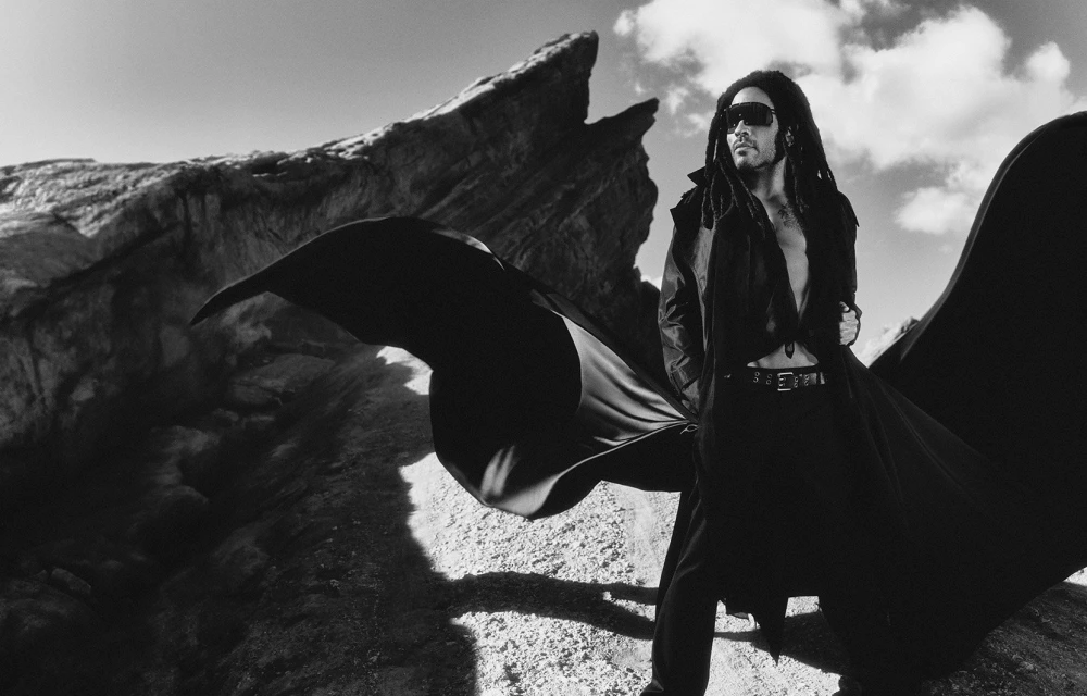 Monochrome outdoor portrait of a dark-haired adult male with dreadlocks wearing sunglasses, a long black coat, standing on a rocky landscape.