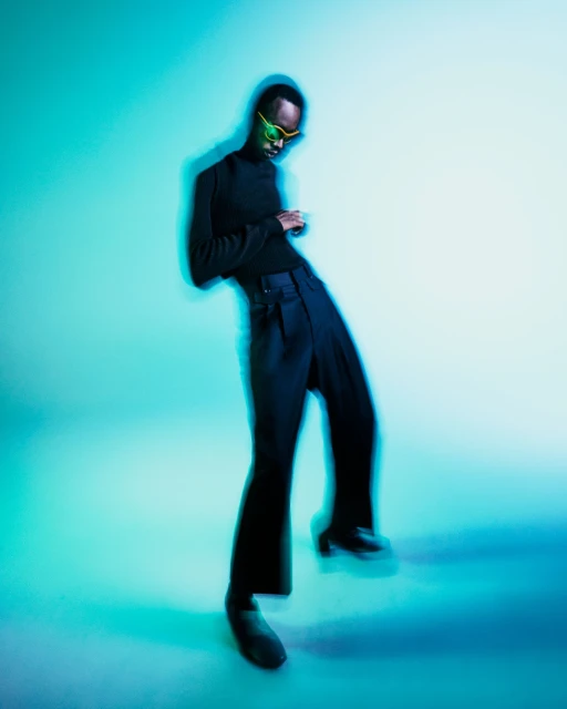 Man in black turtleneck, pants and green sunglasses poses in a studio with cyan lighting against a white and cyan background.