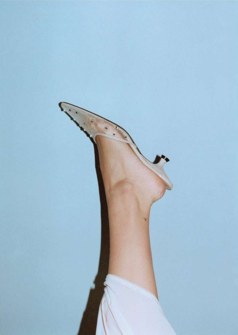 Fashion editorial shot of woman's leg raised with a studded mule shoe against a light blue background in a photo studio setting.