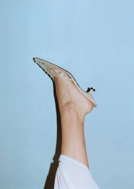 Fashion editorial shot of woman's leg raised with a studded mule shoe against a light blue background in a photo studio setting.