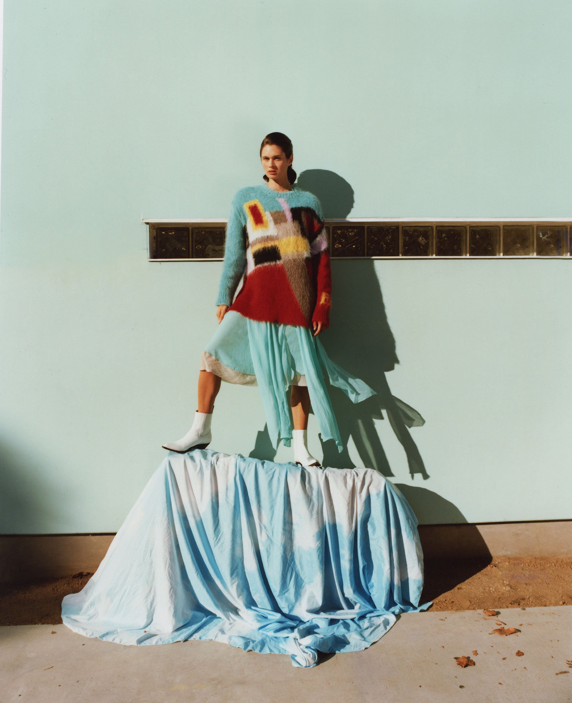 Fashion shot of a woman in an oversized knit sweater and light blue dress posing on a draped surface against a teal wall.