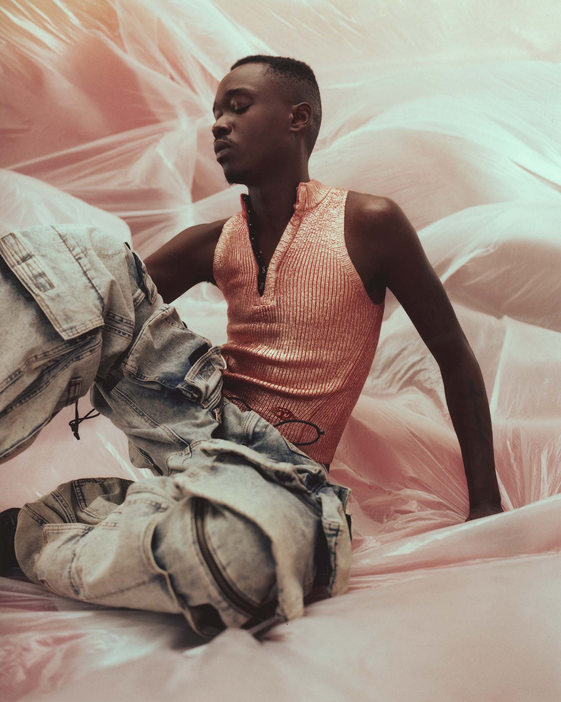 Portrait of a Black man wearing a shiny pink top and patterned pants, sitting amongst pink fabric with eyes closed in a studio setting.