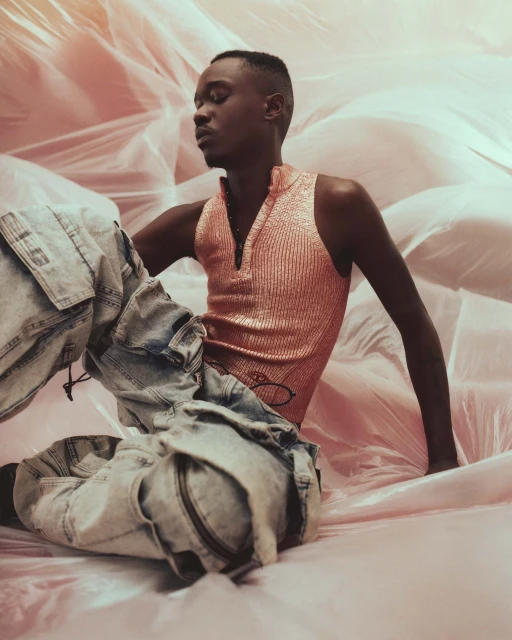 Portrait of a Black man wearing a shiny pink top and patterned pants, sitting amongst pink fabric with eyes closed in a studio setting.