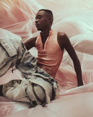 Portrait of a Black man wearing a shiny pink top and patterned pants, sitting amongst pink fabric with eyes closed in a studio setting.