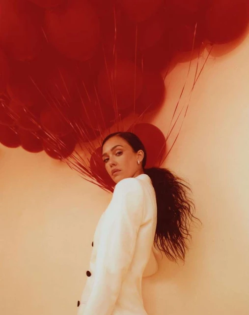 Portrait of a woman in a white blazer with red balloons. The photo is a medium shot taken indoors against a neutral background.