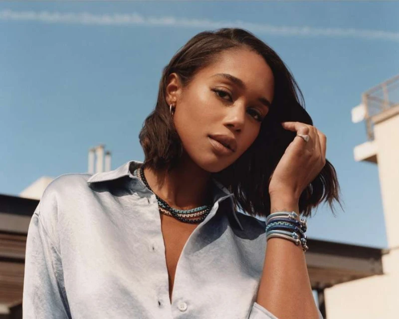 Portrait of a woman wearing jewelry. She has her hand near her head with a bright blue sky and a rooftop in the background.