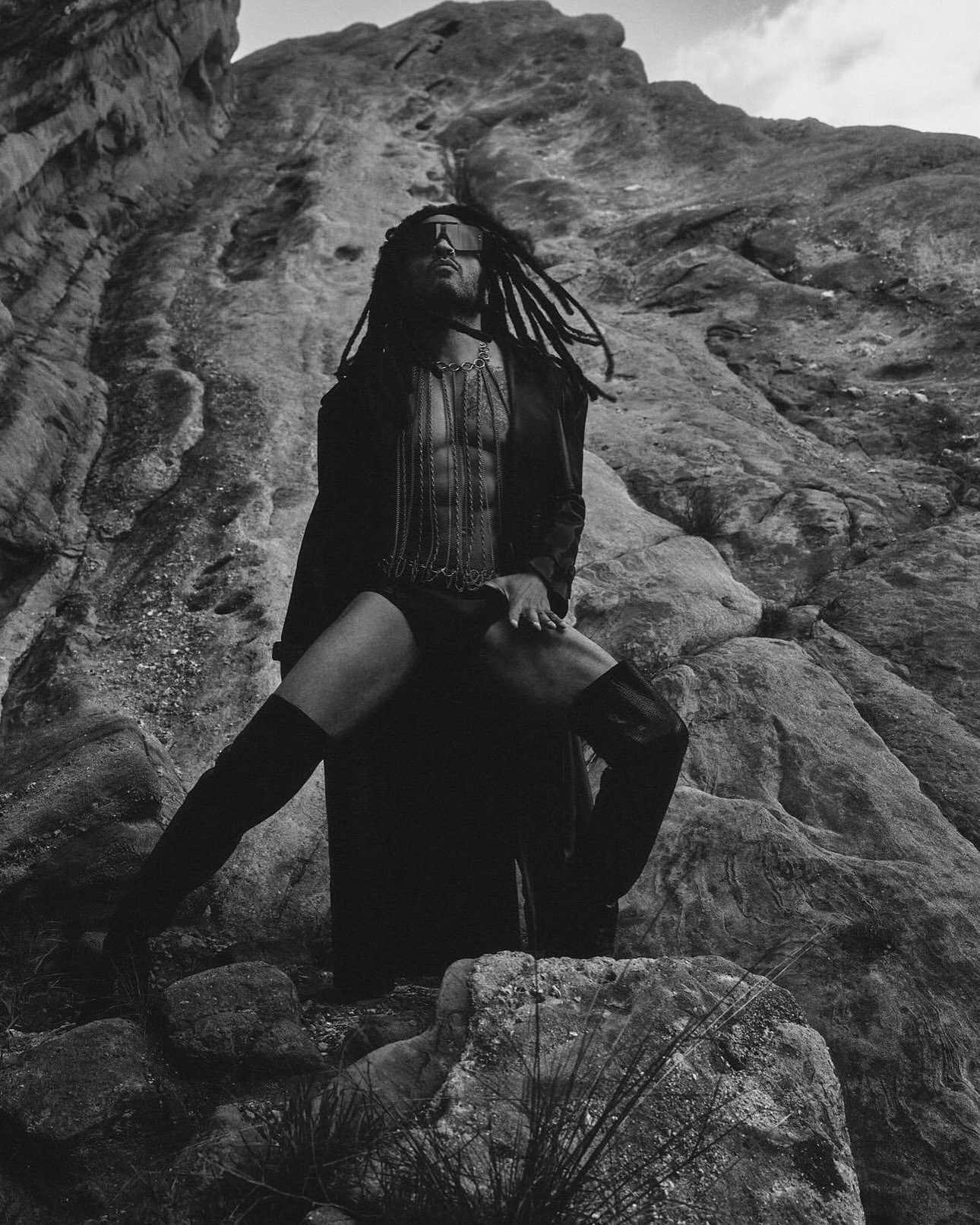 Monochrome portrait of a person with dreadlocks in an open coat, chain top, and boots, posing in front of a textured rock formation.