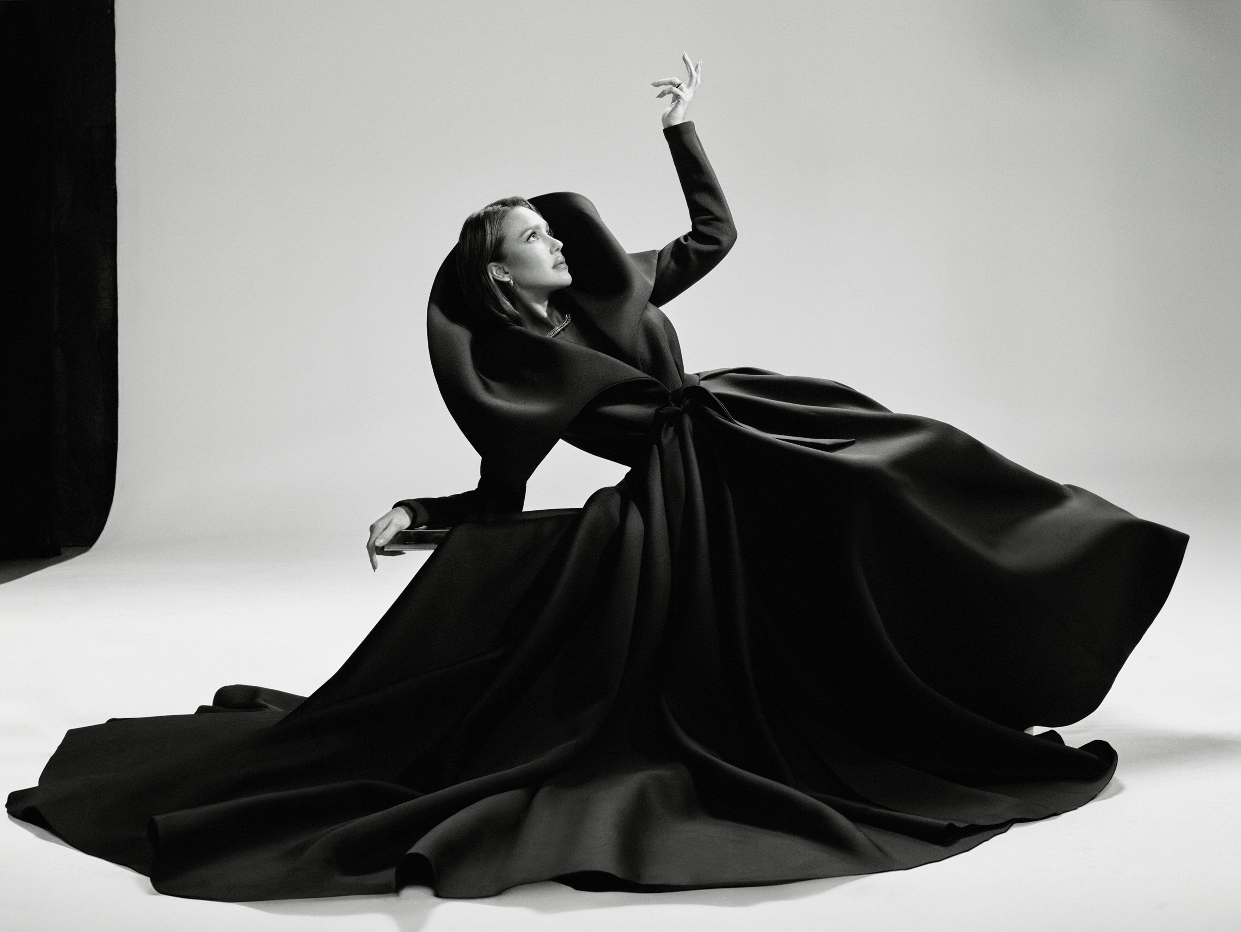 Woman in a dramatic black couture dress sits in a photo studio, posing with one arm raised against a plain background in black and white.