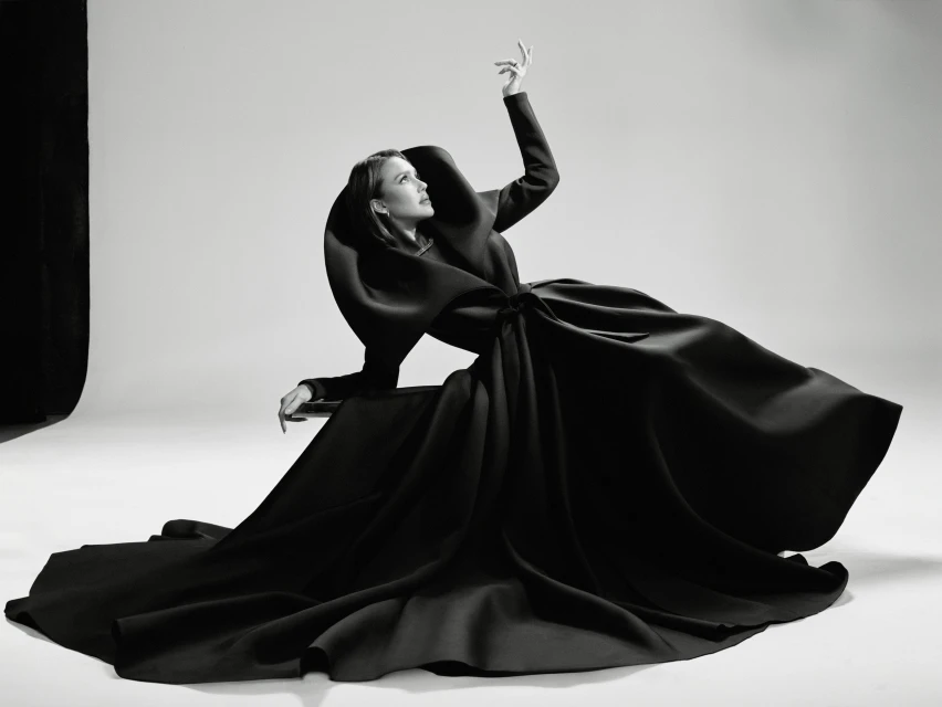Woman in a dramatic black couture dress sits in a photo studio, posing with one arm raised against a plain background in black and white.