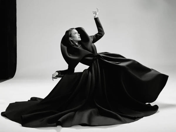 Woman in a dramatic black couture dress sits in a photo studio, posing with one arm raised against a plain background in black and white.
