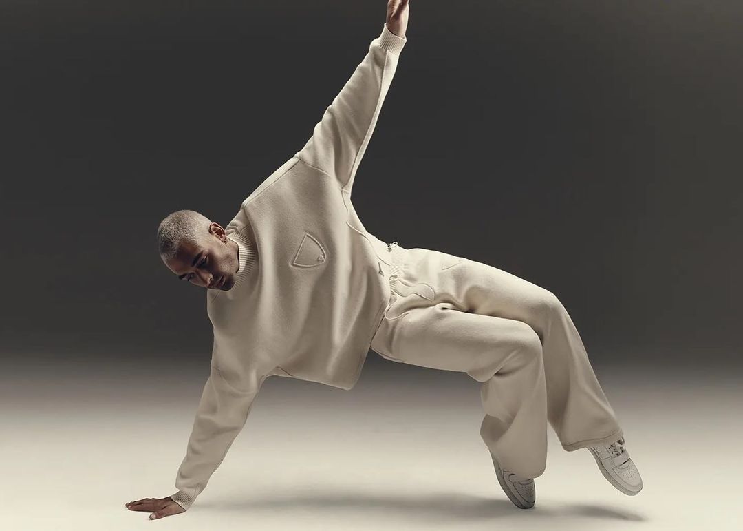An adult male dancer with short gray hair is balanced on one arm in a cream-colored sweater and pants against a neutral gray backdrop.