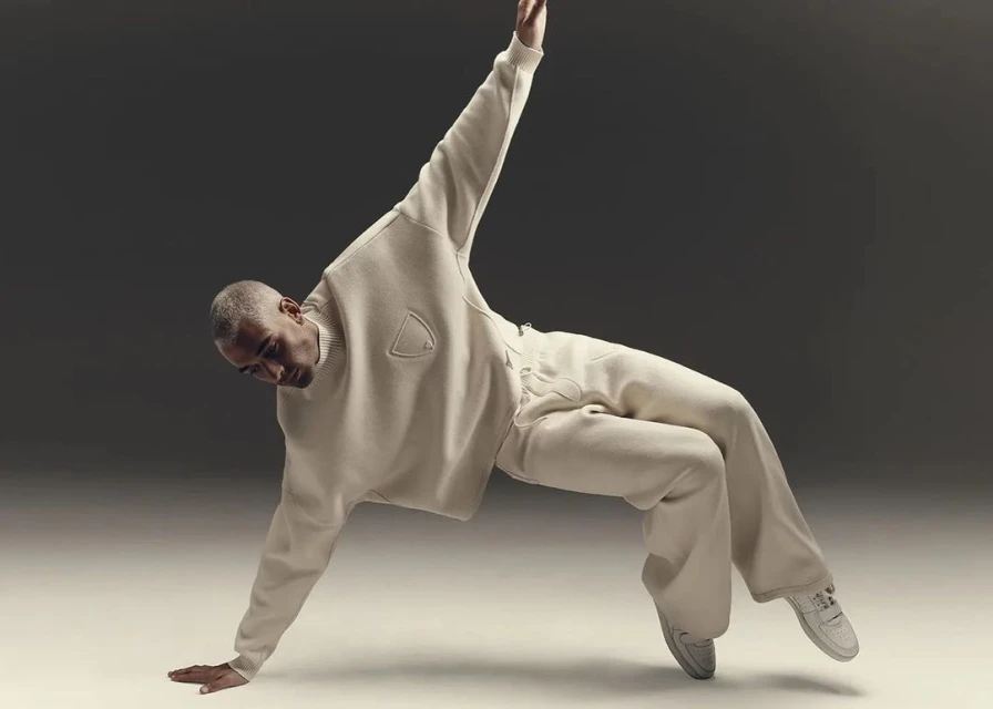An adult male dancer with short gray hair is balanced on one arm in a cream-colored sweater and pants against a neutral gray backdrop.