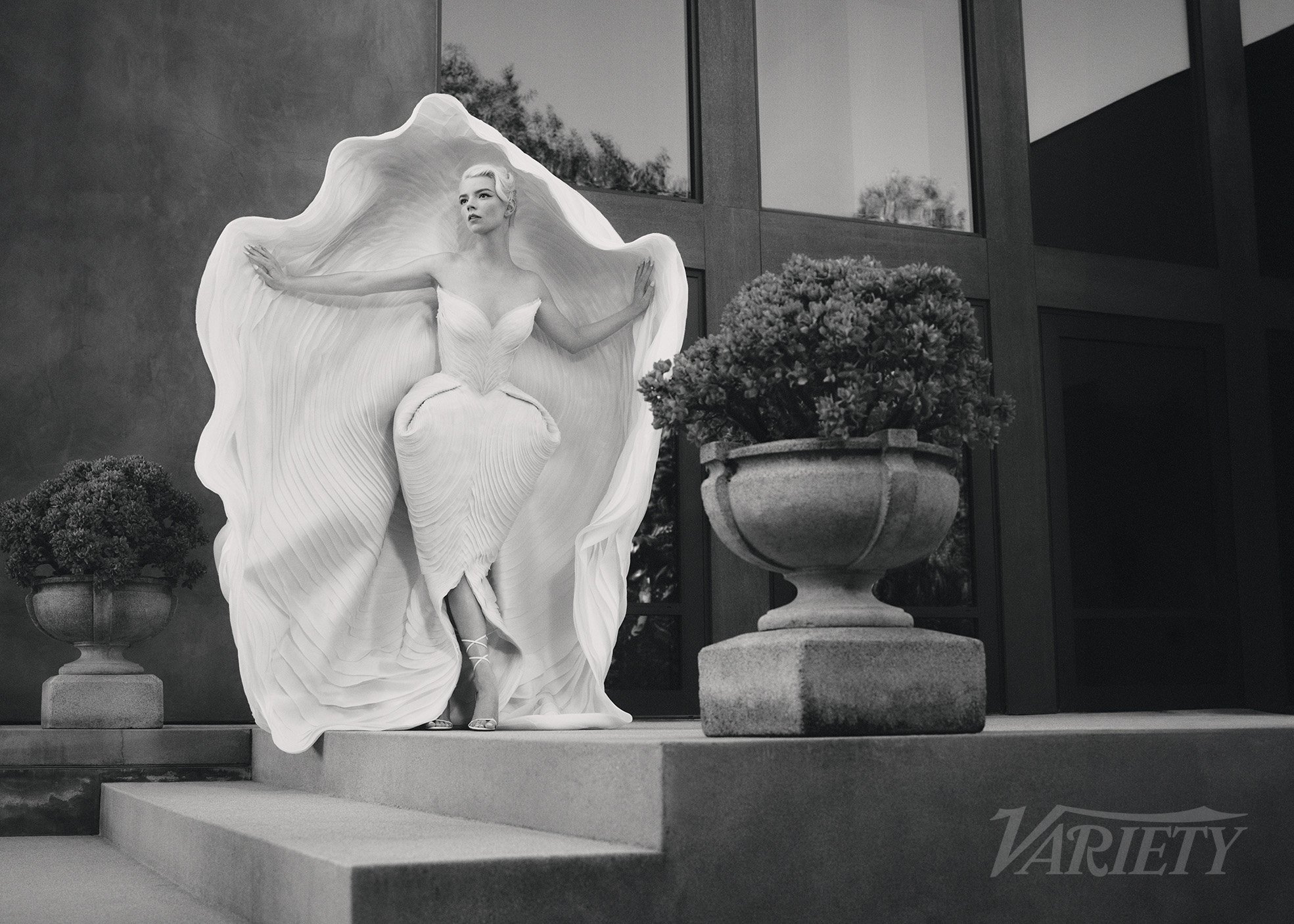 Monochrome portrait of woman in sculpted dress standing on steps with planters in garden, editorial fashion photography.