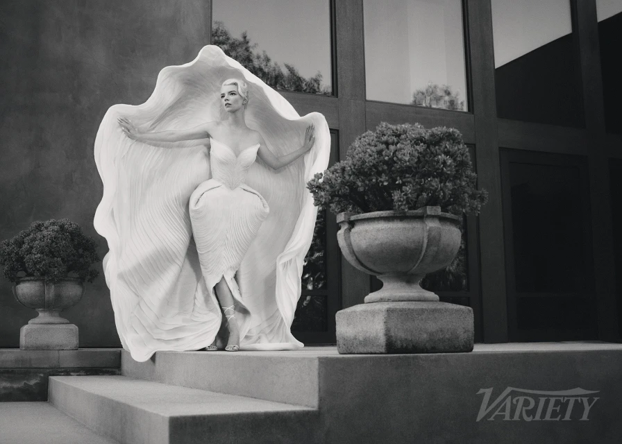 Monochrome portrait of woman in sculpted dress standing on steps with planters in garden, editorial fashion photography.
