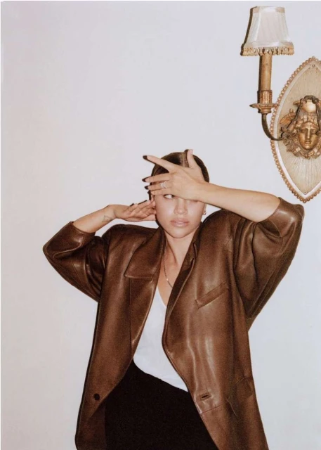 A woman in a brown blazer poses indoors with her hands near her face in front of a wall with an ornate sconce.