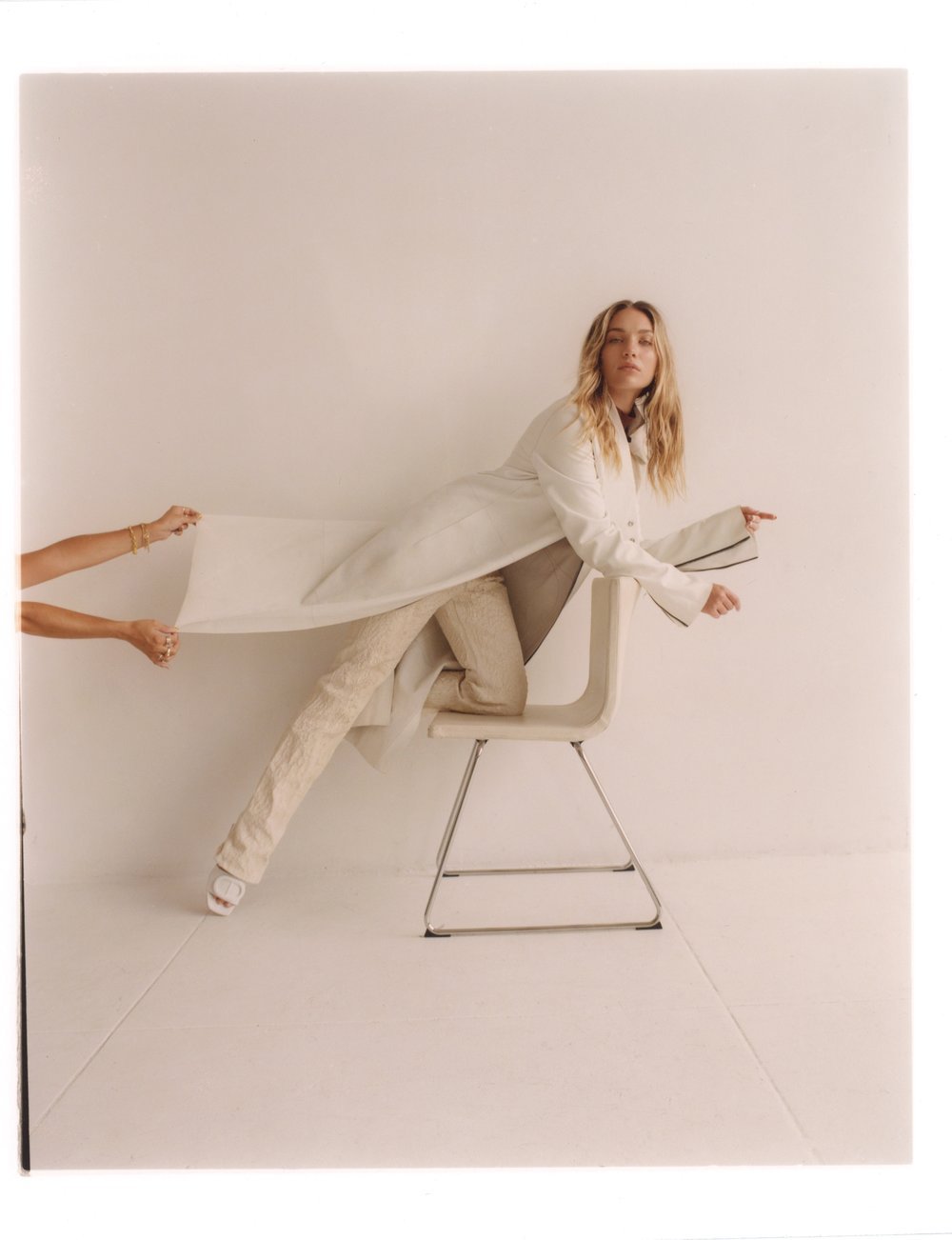 Woman in cream coat and trousers posed dynamically on a white chair while hands pull the coat fabric