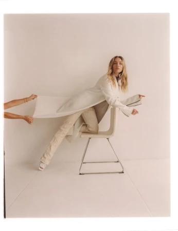 Woman in cream coat and trousers posed dynamically on a white chair while hands pull the coat fabric