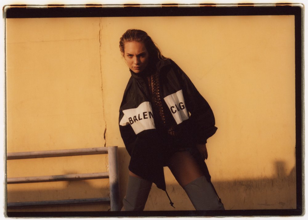 Fashion shot of a woman with wet hair wearing a Balenciaga jacket and grey boots, posing against a yellow wall outdoors.