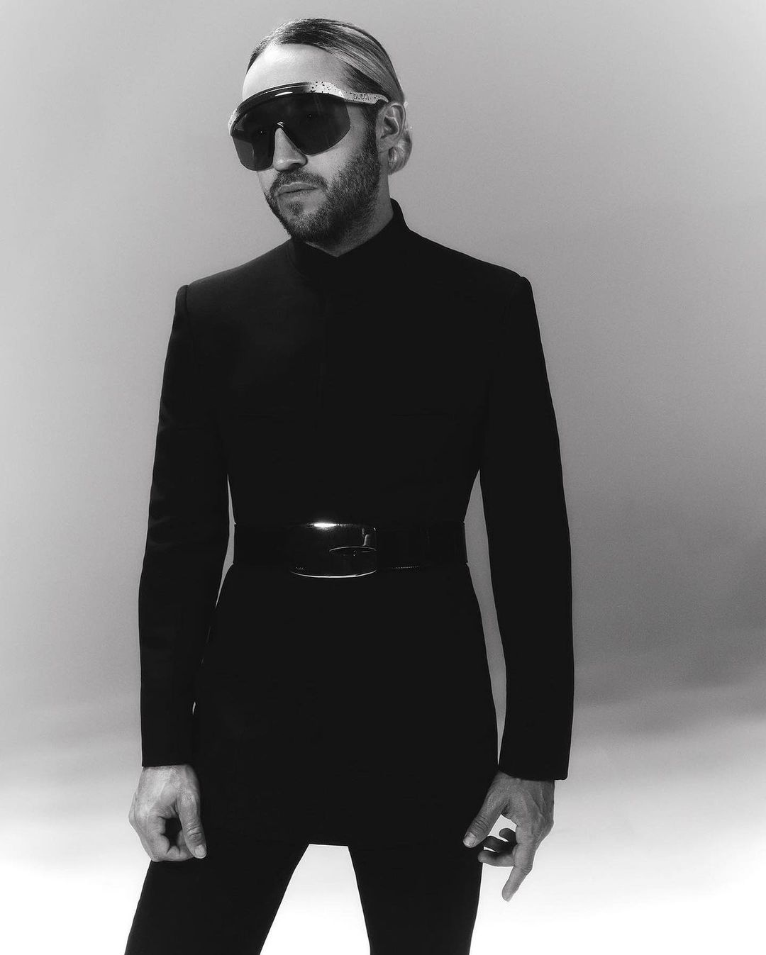Black and white portrait of a man wearing a black bodysuit and sunglasses, standing against a grey background in a photo studio.
