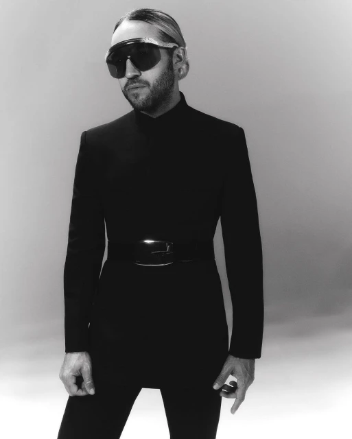 Black and white portrait of a man wearing a black bodysuit and sunglasses, standing against a grey background in a photo studio.