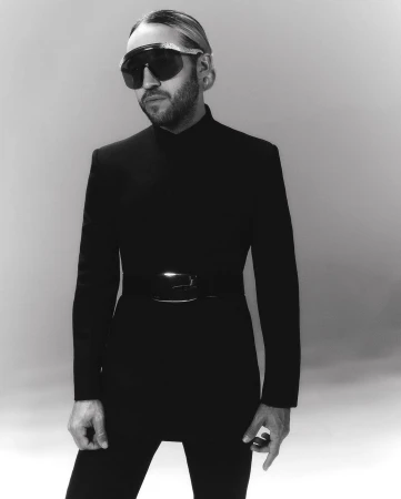 Black and white portrait of a man wearing a black bodysuit and sunglasses, standing against a grey background in a photo studio.