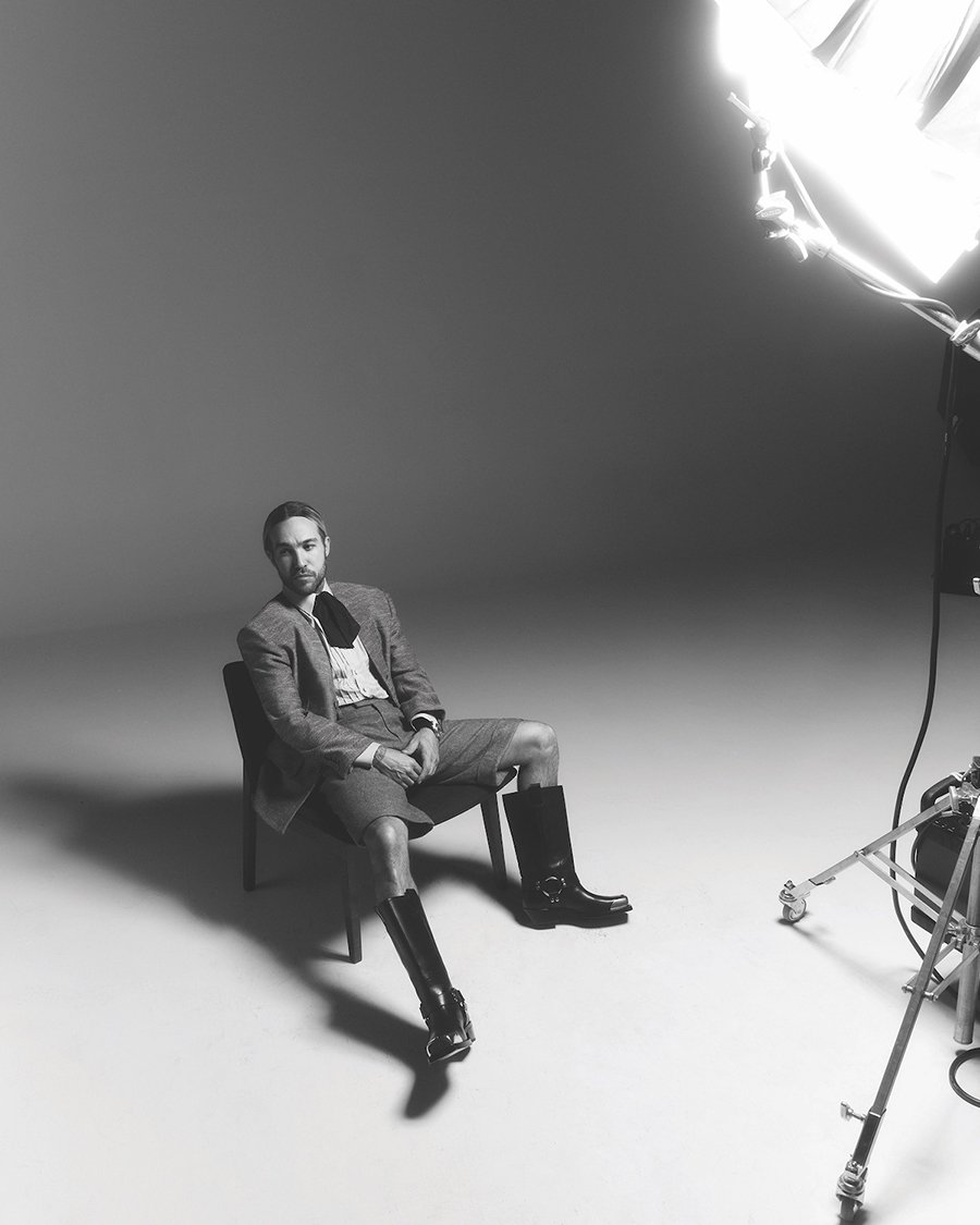 Monochrome portrait of a man in a blazer, shorts, shirt, and boots sitting on a chair in a photo studio under studio lighting.