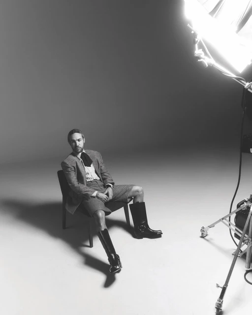Monochrome portrait of a man in a blazer, shorts, shirt, and boots sitting on a chair in a photo studio under studio lighting.