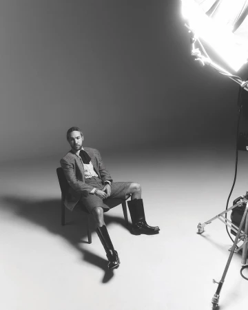 Monochrome portrait of a man in a blazer, shorts, shirt, and boots sitting on a chair in a photo studio under studio lighting.