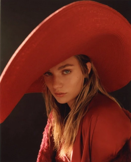Woman in oversized red hat and matching red silky robe poses against a dark background in soft studio lighting.