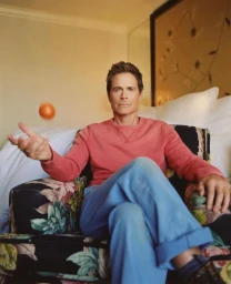 Rob Lowe seated in a floral armchair wearing a pink sweatshirt and blue pants while tossing an orange