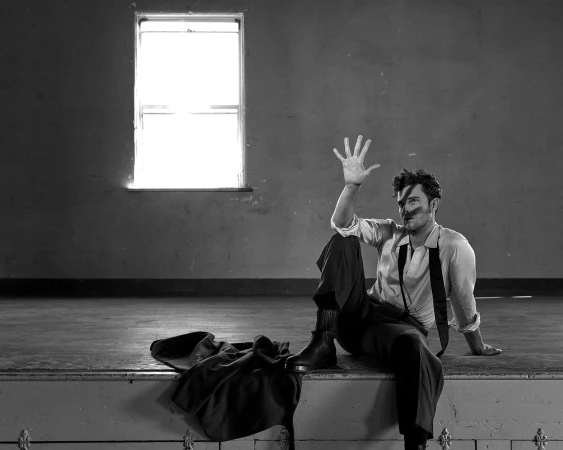 Man in white shirt sitting on stage raising hand with jacket on floor by Jonny Marlow