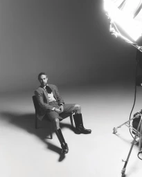 Monochrome studio portrait of Pete Wentz seated on a chair wearing shorts suit and tall boots with a studio light visible
