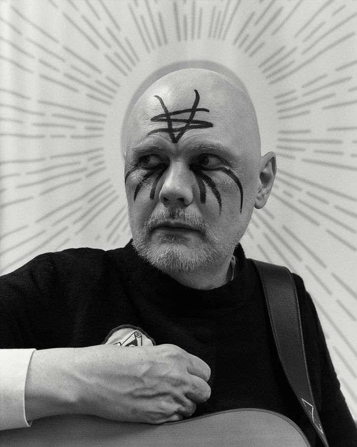 Portrait of Billy Corgan with black face paint markings wearing a black top holding an acoustic guitar by Jonny Marlow