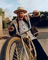 Bryan Cranston wearing a tan fedora and shearling jacket riding a bicycle toward the camera, photographed by Jonny Marlow