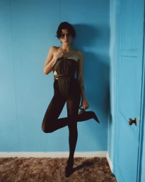 Woman in strapless pleated top and black tights holding a high heel shoe against a blue wall by Jonny Marlow