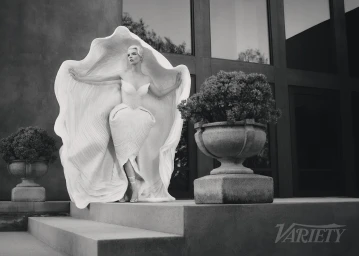 Anya Taylor-Joy wearing a large sculptural white shell dress, posed on stone steps between large urn planters by Jonny Marlow.