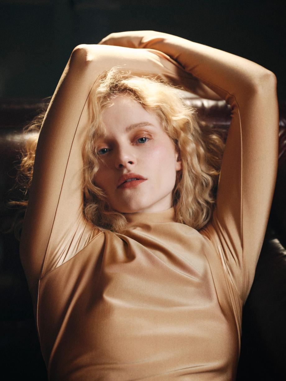 Portrait of a blond-haired woman in a bronze-colored, long-sleeved top, lying on a brown leather couch with arms raised behind her head.