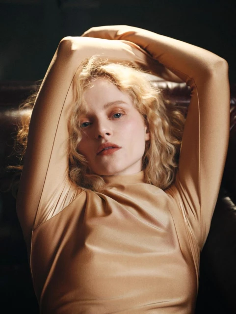 Portrait of a blond-haired woman in a bronze-colored, long-sleeved top, lying on a brown leather couch with arms raised behind her head.