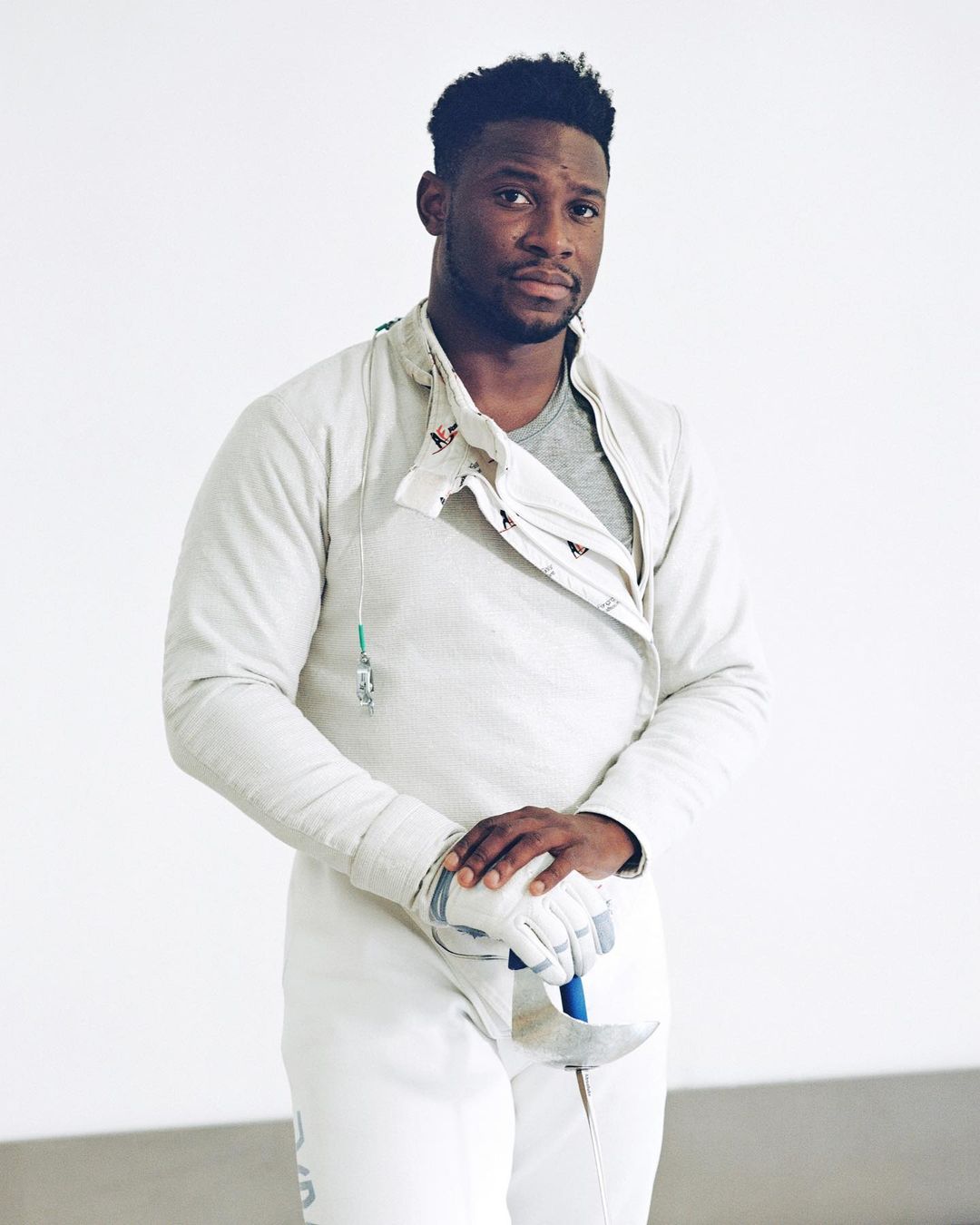 Man in fencing uniform and gloves holding foil indoors. Athletic male in white protective gear ready for sport. 