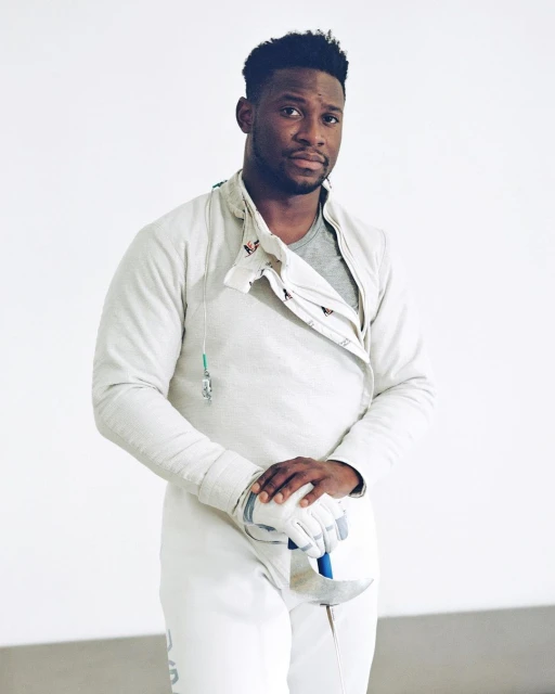 Man in fencing uniform and gloves holding foil indoors. Athletic male in white protective gear ready for sport. 