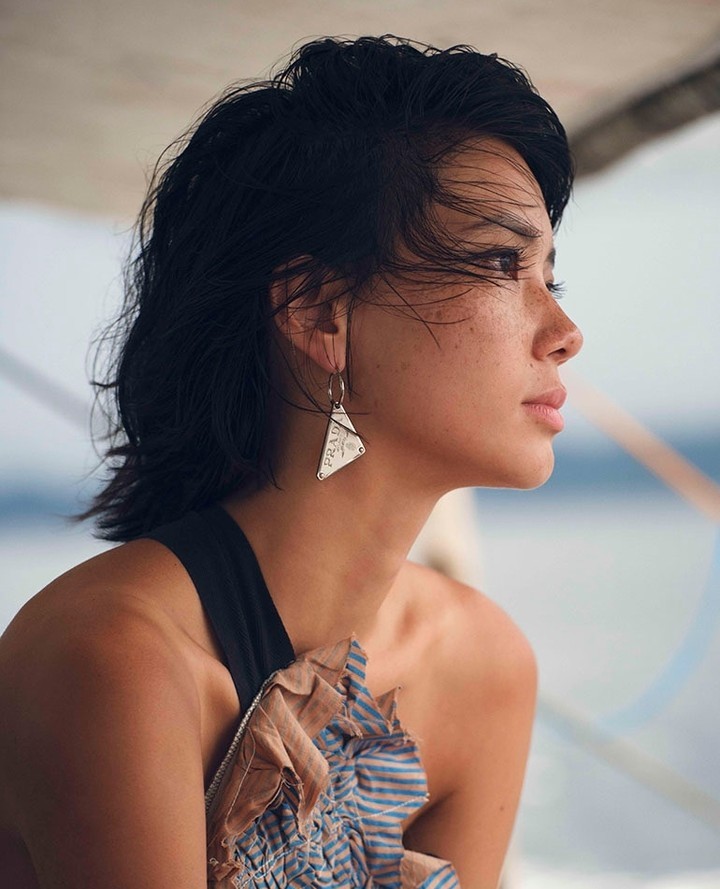 Portrait of a beautiful woman with short dark hair wearing a modern earring, a black tank top, and a blue and beige top, ocean background.