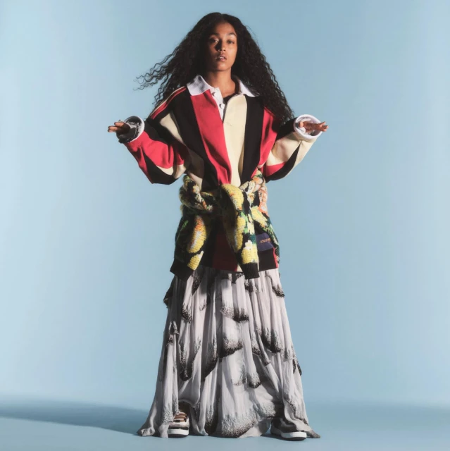 Full-length portrait of a fashionable African-American woman in a striped shirt, long skirt, and sneakers, set against a pale blue backdrop.